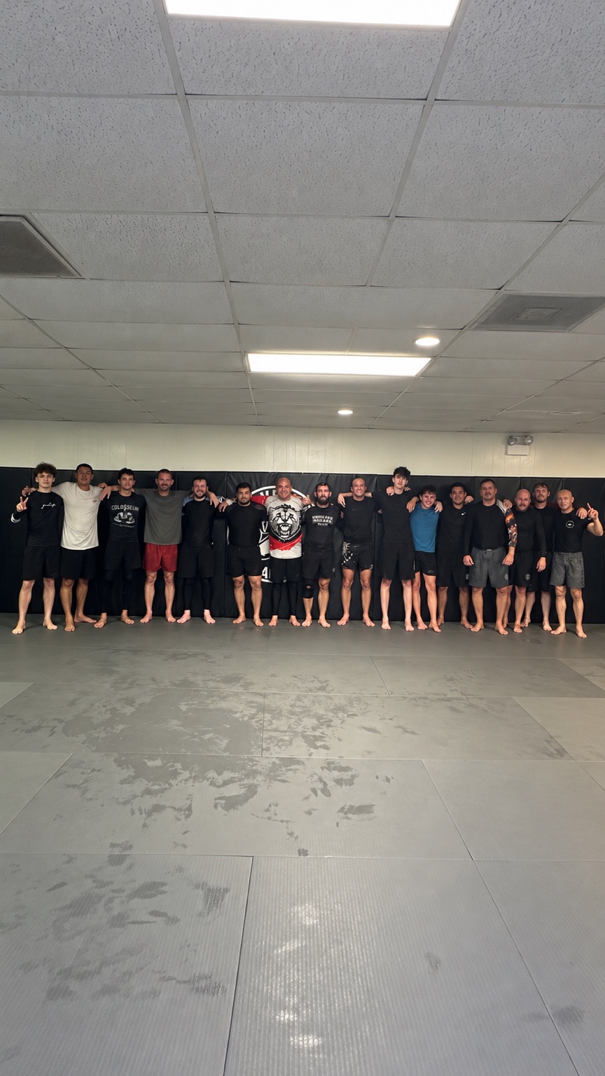 A group of no-gi training partners standing shoulder to shoulder across the academy mat