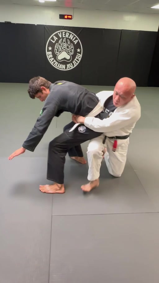 Two gi-clad training partners practicing a standing-to-ground transition at the academy